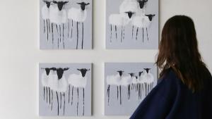 A person in a dark, oversized sweatshirt stands facing a wall with four square canvases. Each canvas, part of the Sunday Selects collection, depicts a minimalistic painting of sheep with black heads and legs on a grey background