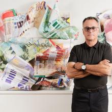 A man stands in a black polo shirt and glasses stands with his arms crossed in front of a large, abstract artwork composed of various colorful banknotes, reflecting the diverse culture celebrated by contemporary artists. 