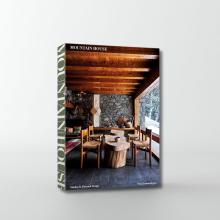A book titled "Mountain House" by Nina Freudenberger features a rustic dining area with wooden beams on the ceiling, a stone wall, and a large wooden table with chairs. The cozy scene includes a window showing a snowy outside view.  