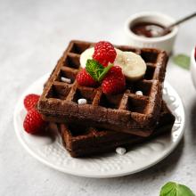 A plate of two Belgium waffles topped with banana slices, raspberries, and a mint leaf. Surrounding the waffles are more raspberries, mini marshmallows, and a small bowl of chocolate sauce. Perfect for brunch on a light-colored table with a white plate. 