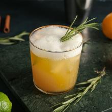 A frothy yellow cocktail, part of our Sunday Selects, is garnished with a sprig of rosemary in a clear glass. Set on a dark green textured surface, the drink exudes art and culture, accompanied by additional rosemary sprigs, a cinnamon stick, and lime in. 