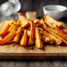 A wooden board topped with freshly made sweet potato wedges garnished with parsley. A salt shaker and a small white dish with dipping sauce can be seen in the background on the dark, rustic table, capturing the essence of culinary art. 
