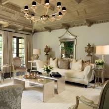 A luxurious living room features a cream-colored sofa with beige and gold pillows, flanked by side tables holding lamps and flowers. An ornate mirror hangs above the sofa, with decorative vases on either side.  