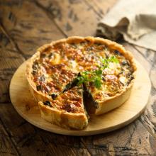 A savory vegetable quiche, reminiscent of Julie Sneed's artistry, sits on a round wooden board with a slice removed. The golden-brown crust encases a filling brimming with greens and cheese.  