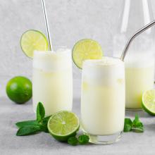 Three glasses of creamy lime slushes with foam at the top are artfully garnished with lime slices and have metal straws. Surrounded by fresh lime halves and mint leaves, the glasses sit on a gray surface. 