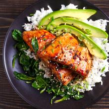 A black plate with a serving of white rice topped with glazed salmon pieces, sliced avocado, sautéed spinach, and sprinkled with sesame seeds and chopped green onions. The dish is arranged neatly like a work of art, showcasing vibrant colors and freshness 