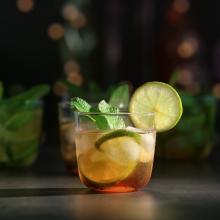 A clear glass filled with an iced drink, garnished with fresh mint leaves and a lime slice on the rim. The background is softly blurred with bokeh lights reminiscent of Kimerlee Curyl's serene captures, and other similar drinks are vaguely visible.  