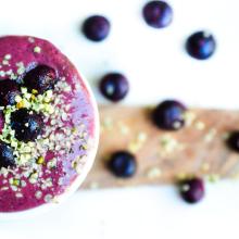 A bowl of vibrant purple smoothie topped with blueberries and chia seeds evokes a sense of Sunday Selects. Surrounding the bowl, scattered blueberries and chia seeds create an artistic display on a light wooden surface against a white background. 