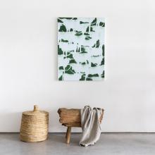 A minimalist interior features a woven basket with a lid on the left, a rustic wooden bench with a beige throw draped over it in the center, and an abstract green and white painting by Julie Sneed hanging on a plain white wall above the bench. The floor i 
