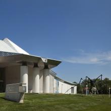 The Kemperr Museum of Art with an angular, white roof and concrete exterior is set against a clear blue sky. A large spider sculpture stands on the grassy lawn in front of the museum, with Louise Bourgeouis spider in front 