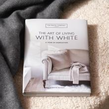 A book titled "The Art of Living with White: A Year of Inspiration" by Chrissie Rucker is placed on a textured, cream-colored surface. The cover shows a cozy white armchair with a matching blanket and cushion.  