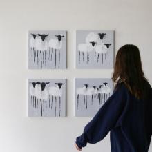 A person in a dark, oversized sweatshirt stands facing a wall with four square canvases. Each canvas, part of the Sunday Selects collection, depicts a minimalistic painting of sheep with black heads and legs on a grey background.  