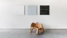 Maura Segal Exhibition Continues A minimalistic room features three abstract paintings on the wall, the left in white, the middle in light blue, and the right in black, each with geometric shapes. 