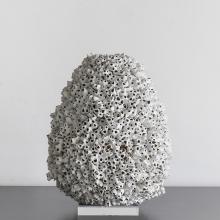 A sculptural piece resembling a large, porous rock or coral reef structure, gray in color and covered with numerous small holes and cavities, sits against a plain white background. The base of the sculpture is flat and appears to rest on a pedestal. This  