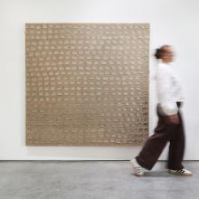 A person wearing a white shirt, brown pants, and sneakers walks past a large, abstract wall artwork. This captivating piece of art features a grid of repetitive, irregular circular shapes on a textured, light brown canvas.  