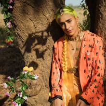 Madame Ghandi in an orange jacket by a tree with flowers next to her 
