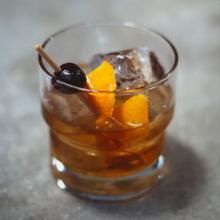 A glass of Old Fashioned cocktail with large ice cubes, garnished with an orange twist and a dark cherry on a cocktail pick, set on a gray surface, evokes the elegance of a Vieux Carre. 