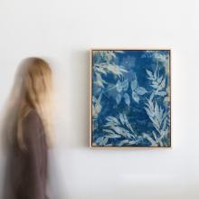 Someday Soon by Kristen Abbott A person with long blond hair, wearing a gray top, observes a framed cyanotype on a white wall. The artwork features a blue background with white botanical patterns, resembling leaves and flowers. Slightly blurred, the person suggests movement, adding life to Kristen Abbott's serene silk artwork.