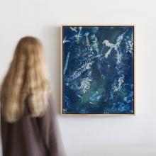 Time Will Tell by Kristen Abbott A person with long blond hair stands admiring a large abstract painting dominated by blue and white colors on a white wall. Created by a mixed media artist, the painting features swirling patterns that evoke movement, reminiscent of the cyanotype process.