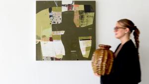 Woman holding a ceramic pot, standing in front of a vibrant abstract collage artwork by Deborah T. Colter. The artwork features bold geometric shapes and a mix of warm and cool colors."